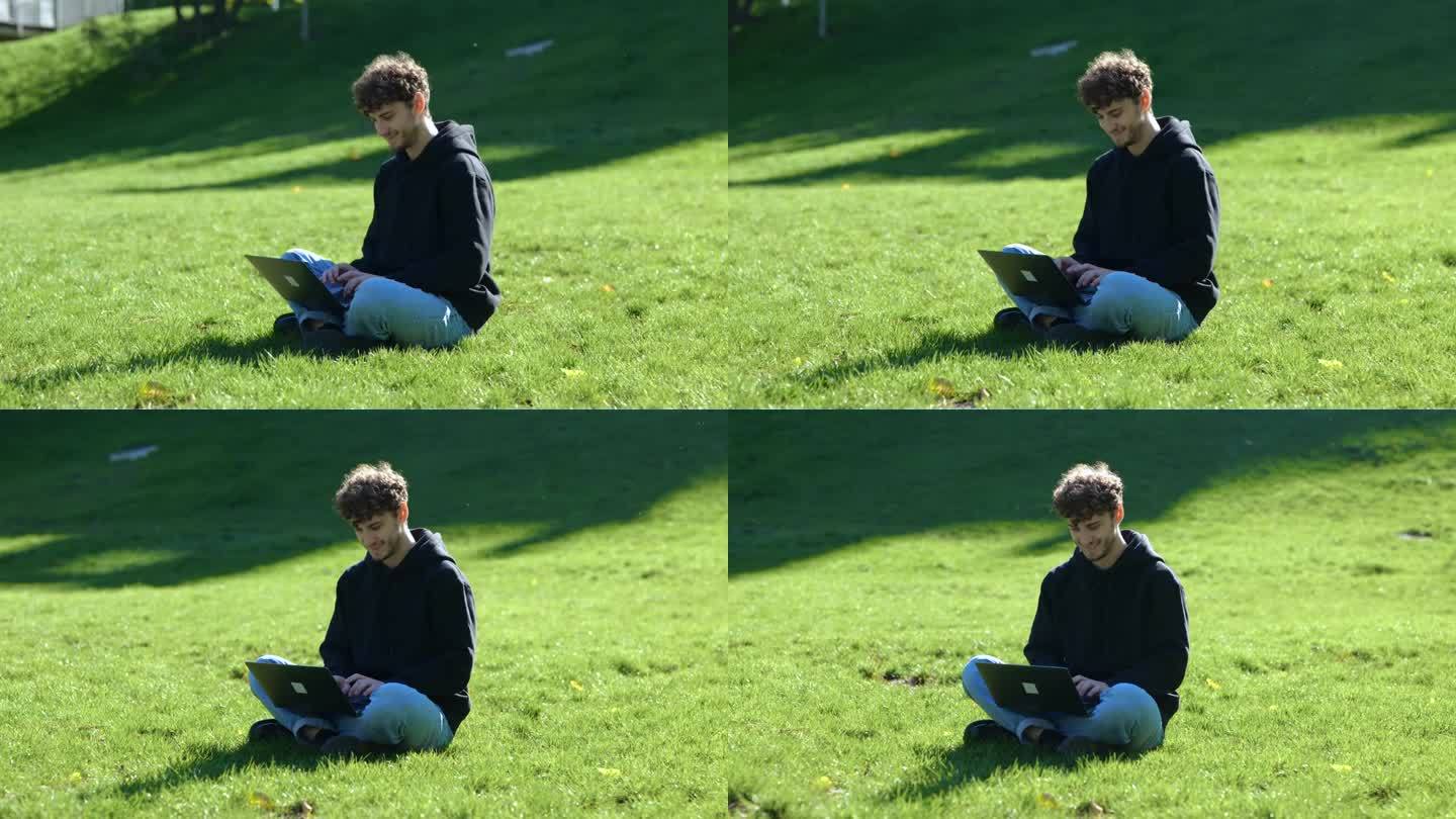 微笑的年轻人坐在公园的草地上，一边用笔记本电脑打字一边工作。秋天的季节。教育，工作理念。慢动作