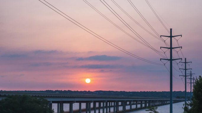 红黄色的太阳球从横跨河流的公路桥上的电线杆后面的薄云中升起，时间流逝