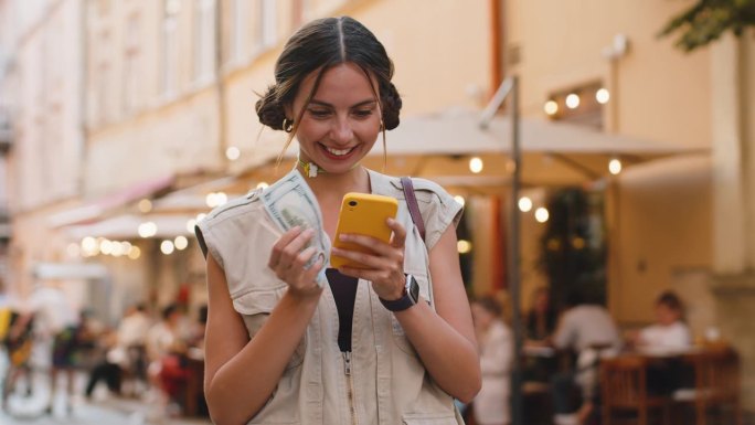 快乐的年轻女子在城市街道上用智能手机计算应用程序数钱