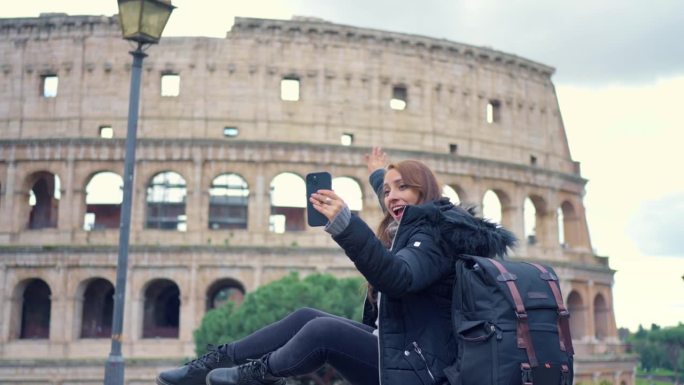 快乐的年轻拉丁女游客带着行李和保暖的衣服，在意大利罗马的历史建筑斗兽场前，在多云的天空中用智能手机记