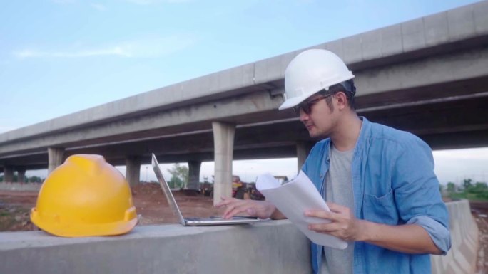 图为，亚洲的土木工程师正在用笔记本电脑和设计图检查在施工现场进行的高速公路工程。