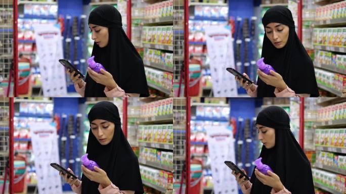 在超市里，戴着头巾、拿着智能手机、拿着一瓶洗发水的女人在网上看化妆品的评论。戴头巾的穆斯林妇女负责地