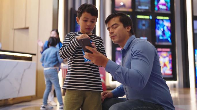 亚洲父亲和小儿子在电影院用智能手机购买电影票。父子俩周末一起在电影院通过智能手机预订座位。家庭活动概