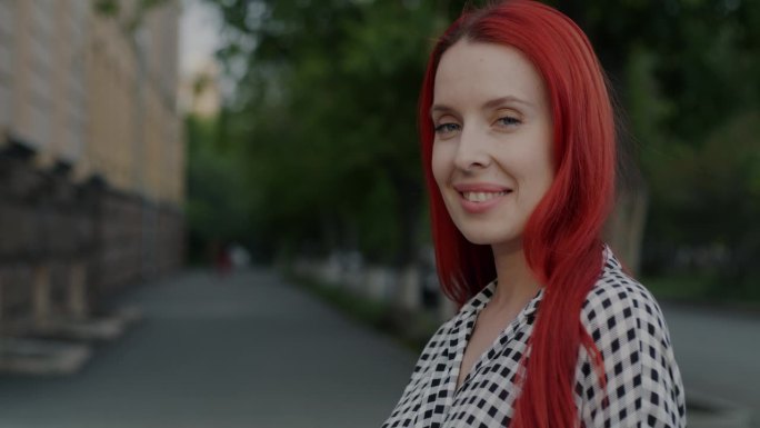 一个染红头发的漂亮年轻女子的慢动作肖像转向镜头，微笑着在城市户外调情