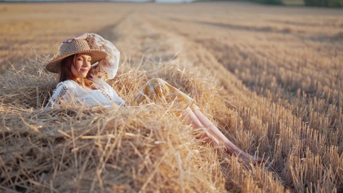 一个漂亮的女人，头戴草帽，身穿绣花上衣，躺在夕阳下乡间的干草上。乡村自然，干草堆，度假，放松和收获的