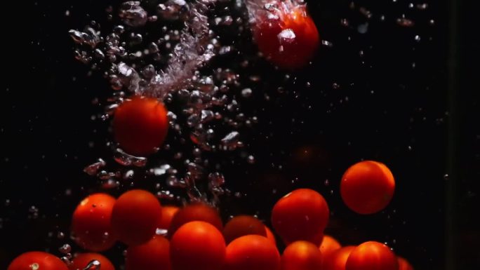 红色成熟的圣女果落在透明的水中，黑色的背景。新鲜的有机蔬菜在鱼缸里飞溅，产生气泡。
