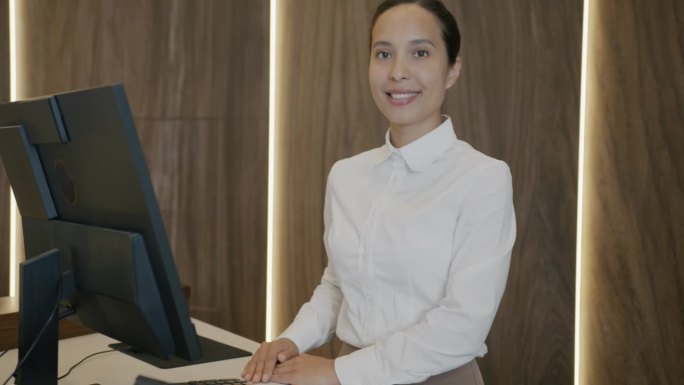年轻的女接待员站在酒店前台，带着电脑微笑