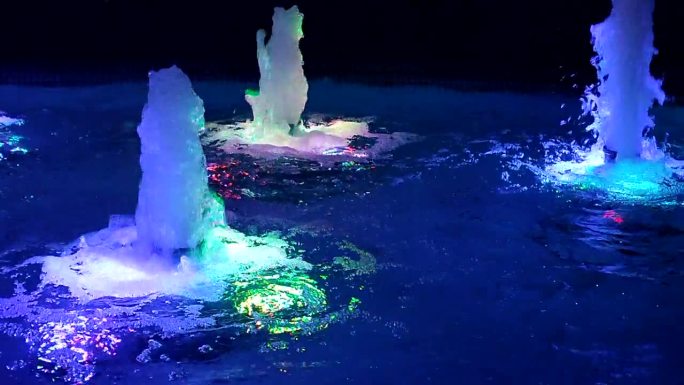 夜晚，五彩缤纷的灯光照亮了水中的喷泉。水流上升，被多色灯光照亮。喷泉喷涌。水压，水头。德科装饰