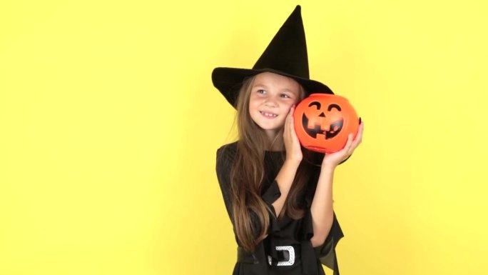 万圣节的概念。肖像微笑的小女孩，戴着黑帽子的女巫，脸上拿着橙色的南瓜，在工作室的黄色背景上孤立地摆姿