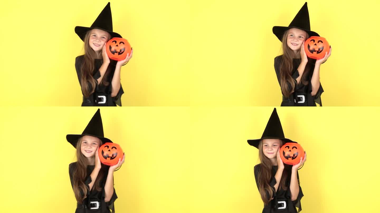万圣节的概念。肖像微笑的小女孩，戴着黑帽子的女巫，脸上拿着橙色的南瓜，在工作室的黄色背景上孤立地摆姿