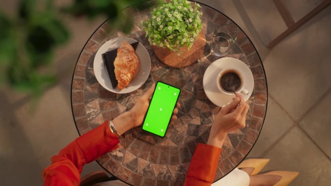 咖啡馆里匿名女性使用智能手机的俯视图，模拟绿屏Chromakey显示屏。女人在放松和喝咖啡的时候查看