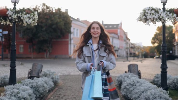 慢镜头迷人的微笑漂亮的年轻女子走在日落时分的城市街道上，带着购物袋，转过身来，对着镜头微笑，对购物和
