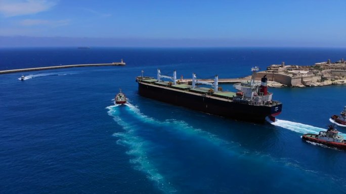 一艘巨型货船在三艘拖船的帮助下离开马耳他瓦莱塔港的独特航拍照片。蓝色开阔的大海和石灰岩建筑作为背景。