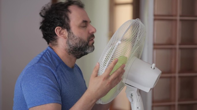 夏日男子在家享受电扇吹来的凉风。一名男子在通风机前酷暑，用电扇冷却器降温。炎热的天气，气候变化。