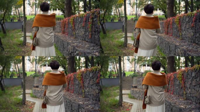 现场摄像机跟随自信的高加索妇女在阴天的秋天公园散步。跟踪拍摄身穿时尚服装、悠闲优雅的女士在户外散步、