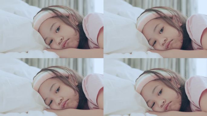 可爱的亚洲女孩的脸部特写，她独自睡在她的床上，白色的毯子和明亮的灯光显示了孩子的无聊，紧张，不快乐，