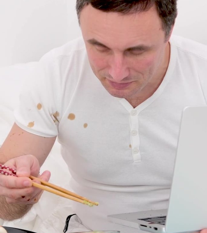 男人坐在白色的床上，手里拿着笔记本电脑，用筷子吃寿司，掉进酱油里，把t恤溅到电脑里的笔记本电脑上，在
