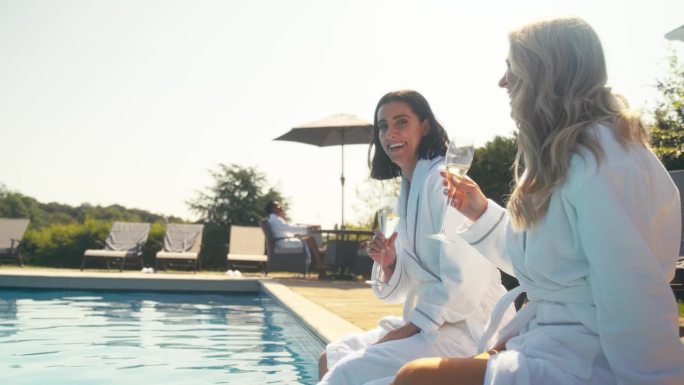 两个成熟的女性朋友穿着长袍，在室外泳池边喝香槟