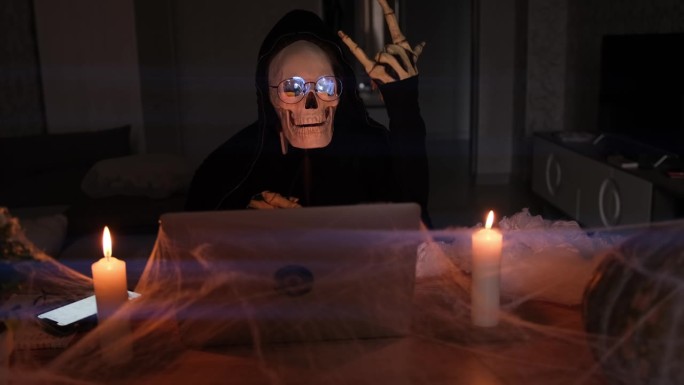 神秘男子在可怕的骷髅服装显示摇滚符号手坐在桌子上的笔记本电脑与蜘蛛网和蜡烛