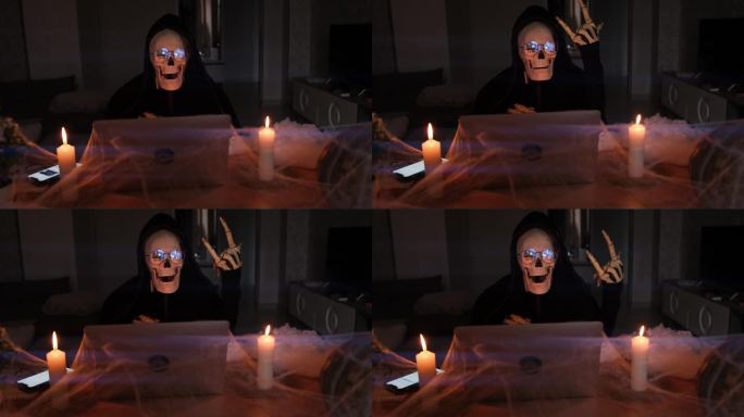 神秘男子在可怕的骷髅服装显示摇滚符号手坐在桌子上的笔记本电脑与蜘蛛网和蜡烛