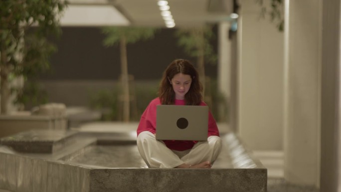 一个年轻的女人坐在莲花式的姿势上，用笔记本电脑打字，酒店大厅里有游泳池