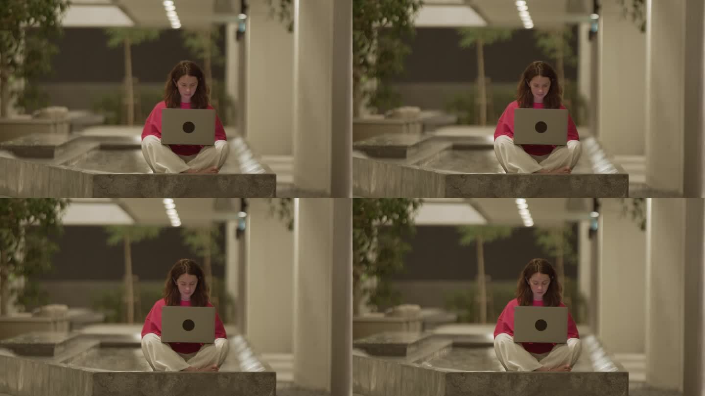 一个年轻的女人坐在莲花式的姿势上，用笔记本电脑打字，酒店大厅里有游泳池