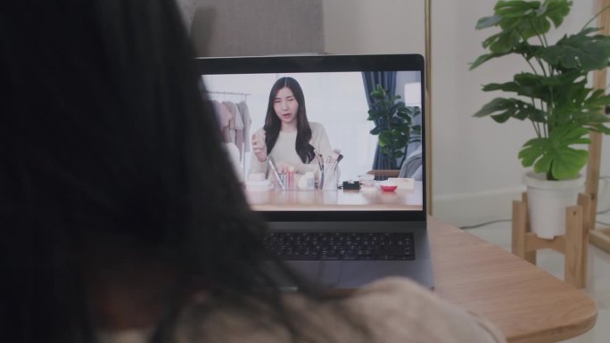 年轻女子正在用笔记本电脑观看美妆博主点评化妆品和化妆教程直播视频。