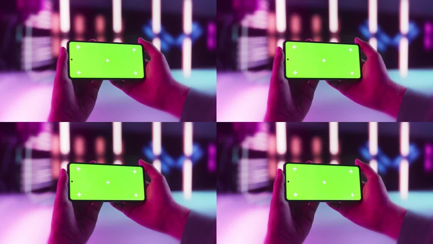 一个时尚的人在霓虹灯工作室的照相机前拿着一部现代手机。智能手机与绿色屏幕色度键显示用于电子学习和教育