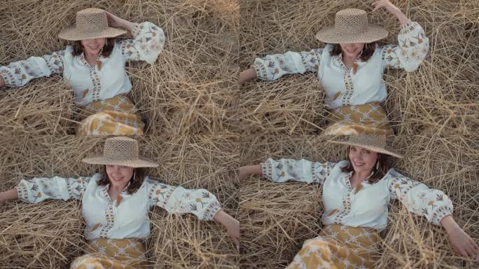 美丽的女人戴着草帽，穿着绣花上衣，微笑地躺在乡村的干草上，夕阳西下。乡村自然，干草堆，度假，放松和收