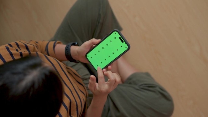 一个女人拿着智能手机，坐在家里的地板上滚动着绿屏模型。