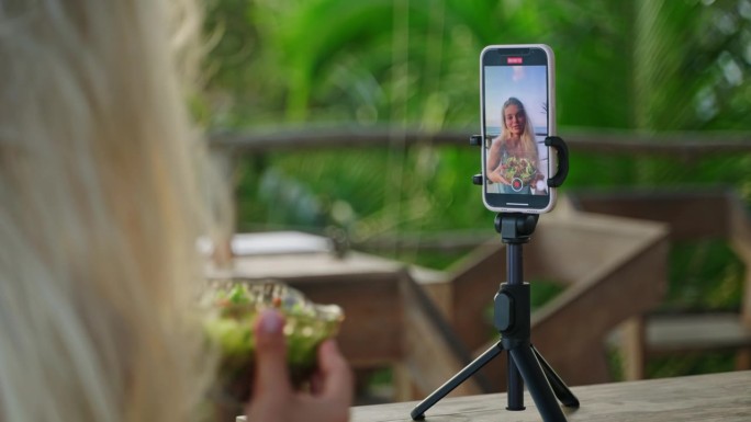 智能手机屏幕上，年轻女博主在热带岛屿的咖啡馆里用蔬菜沙拉自拍视频。三脚架上的手机拍下了女性旅行者在户