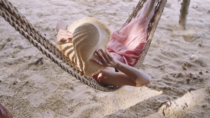一名身穿粉色连衣裙的亚洲女子用草帽遮住脸，躺在吊床上，沙滩的后景