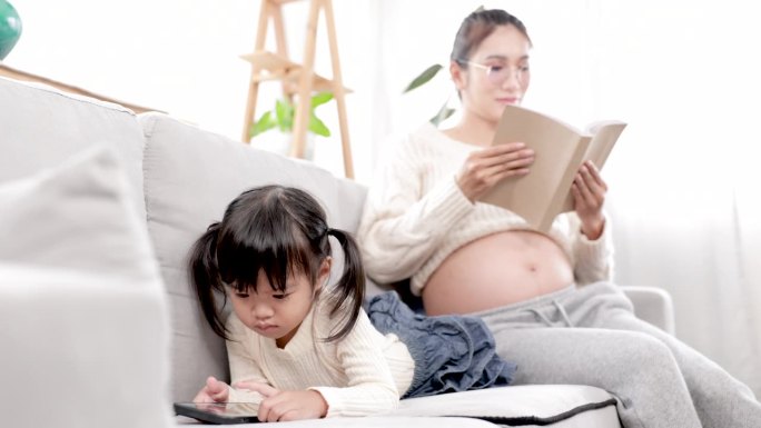 4k，亚洲孕妇妈妈看书而可爱的女儿躺在沙发上看智能手机，妈妈和孩子女孩一起花时间与爱温柔，生活方式怀