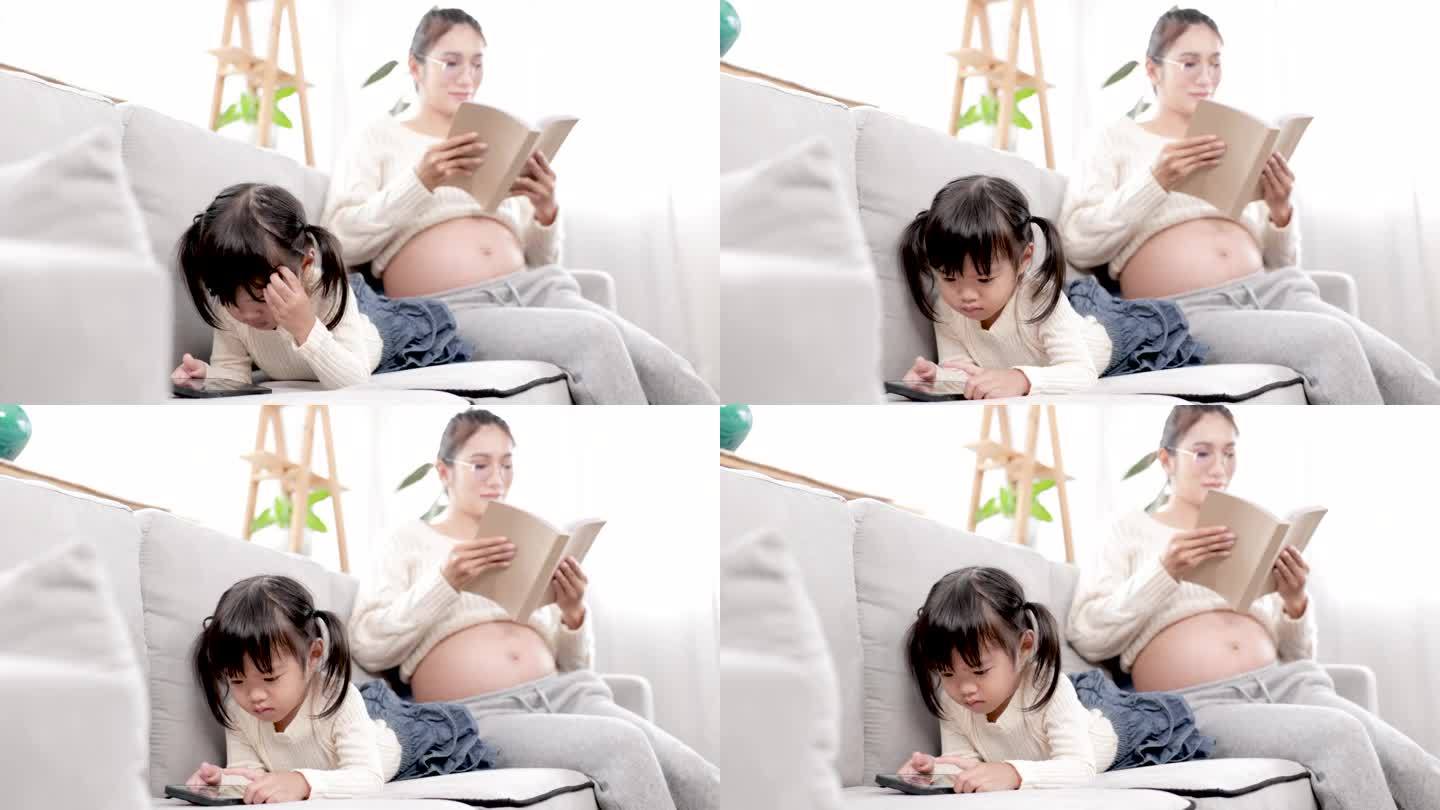 4k，亚洲孕妇妈妈看书而可爱的女儿躺在沙发上看智能手机，妈妈和孩子女孩一起花时间与爱温柔，生活方式怀