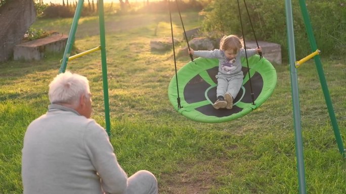 祖父在夏日夕阳下荡秋千。爷爷和孙子坐在公园里的秋千上。60多岁的老爷爷推着漂亮的宝宝玩跷跷板。老人和