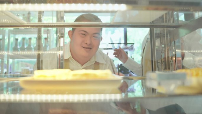 年轻的亚洲残疾男子和他的高级女同事穿着围裙，站在冰箱前，按照顾客在咖啡馆点的面包送到顾客家。残疾咖啡