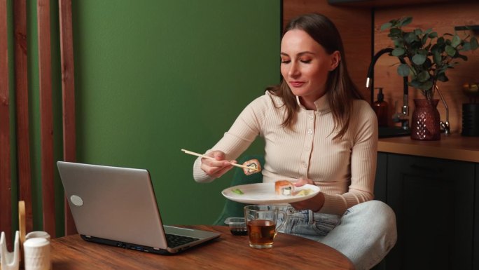 穿着米色运动衫的黑发美女坐在厨房里吃着寿司，用笔记本电脑看电影。女人在周末和节假日休息