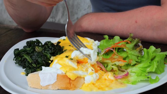 特写镜头男子用叉子扎起一块荷包蛋，蘸上蛋黄。男士早餐在餐厅吃荷包蛋，沙拉配番茄黄瓜洋葱吃荷包蛋可以保