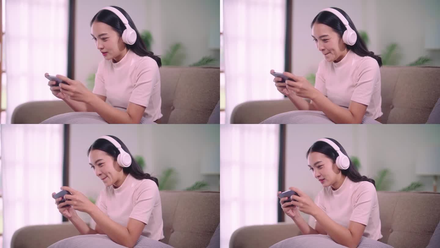兴奋的年轻漂亮的亚洲女人玩手机游戏赢得胜利的时刻，戴上无线耳机，坐在沙发上在她的家客厅的乐趣。