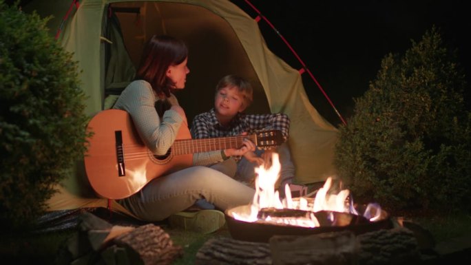 晚上妈妈和孩子坐在营火旁的帐篷里弹吉他唱歌。幸福的一家人在黑暗中围坐在火边唱歌。