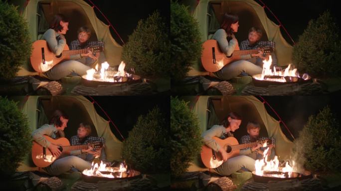 晚上妈妈和孩子坐在营火旁的帐篷里弹吉他唱歌。幸福的一家人在黑暗中围坐在火边唱歌。