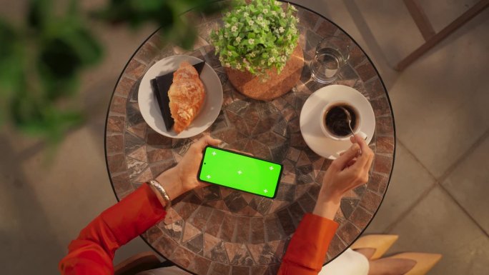 咖啡馆里匿名女性使用智能手机的俯视图，模拟绿屏Chromakey显示屏。一位女士一边放松一边喝咖啡，