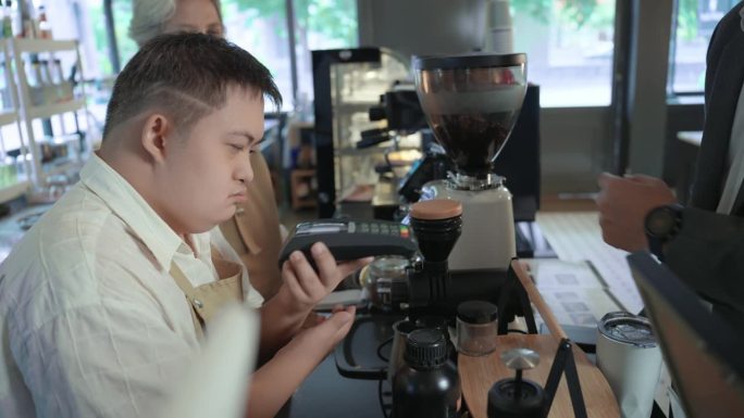 一名患有唐氏综合症的年轻亚洲男子在点完美味的咖啡后，将信用卡机递给了他的顾客。一名残疾青年和他的资深