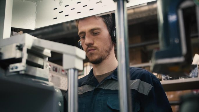 一个红头发的男人戴着耳机在一家服装厂的工业机器上工作的特写镜头。工作日开始在工厂工作