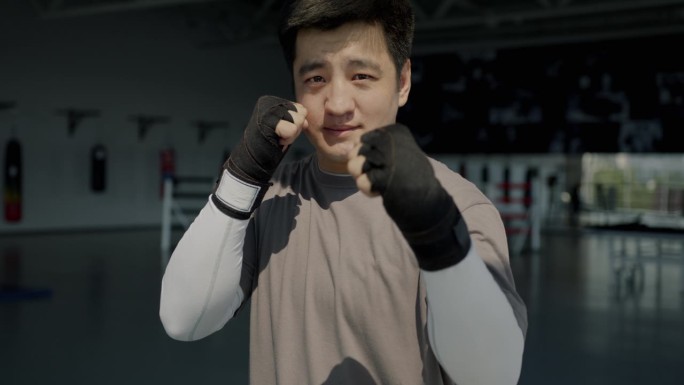自信的亚洲男子综合格斗选手站在健身房，双手包裹着看着摄像机准备战斗的肖像