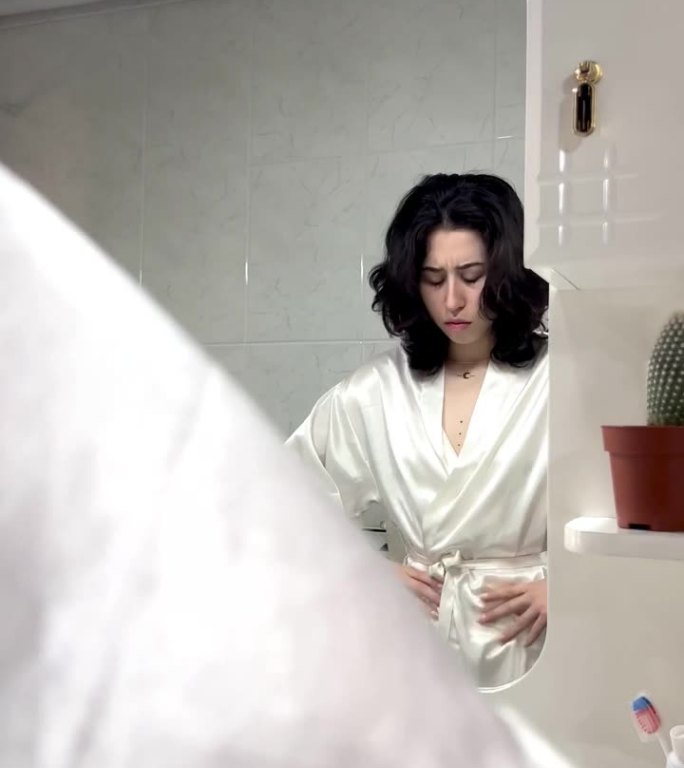 视频中，一位身着长袍的年轻拉丁女性站在家里的镜子前，双手放在肚子上，抱怨自己胃痛。