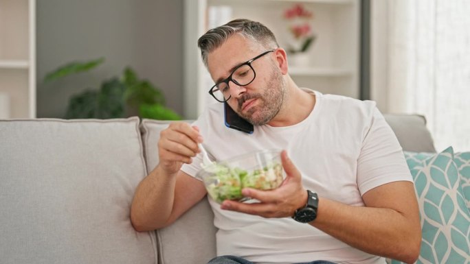 头发花白的男人在家里吃沙拉，用智能手机聊天