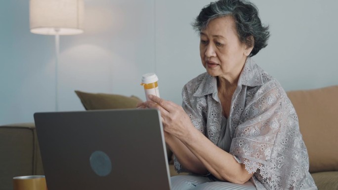 亚洲老年妇女在客厅用电脑，拿着药瓶向医生询问有关药物的问题。