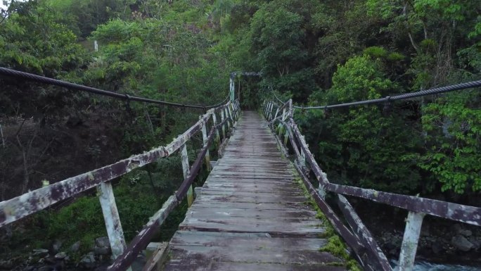 FPV在热带丛林中走过一座破旧的小木桥，桥上有粗金属电缆，下面有一条小河