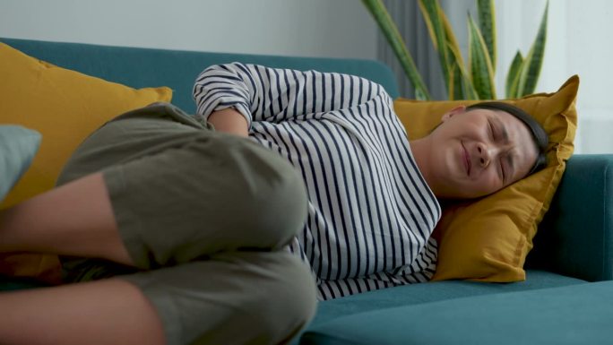 一位亚洲妇女胃痛得躺在家里客厅的沙发上。